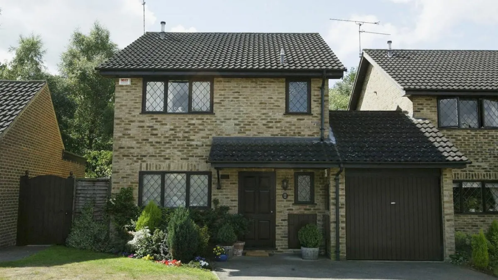 A two-story brick house with a dark tiled roof, featuring multiple windows with diamond-shaped panes, a wooden front door, and a garage door on the right. The front yard is landscaped with various plants and flowers.