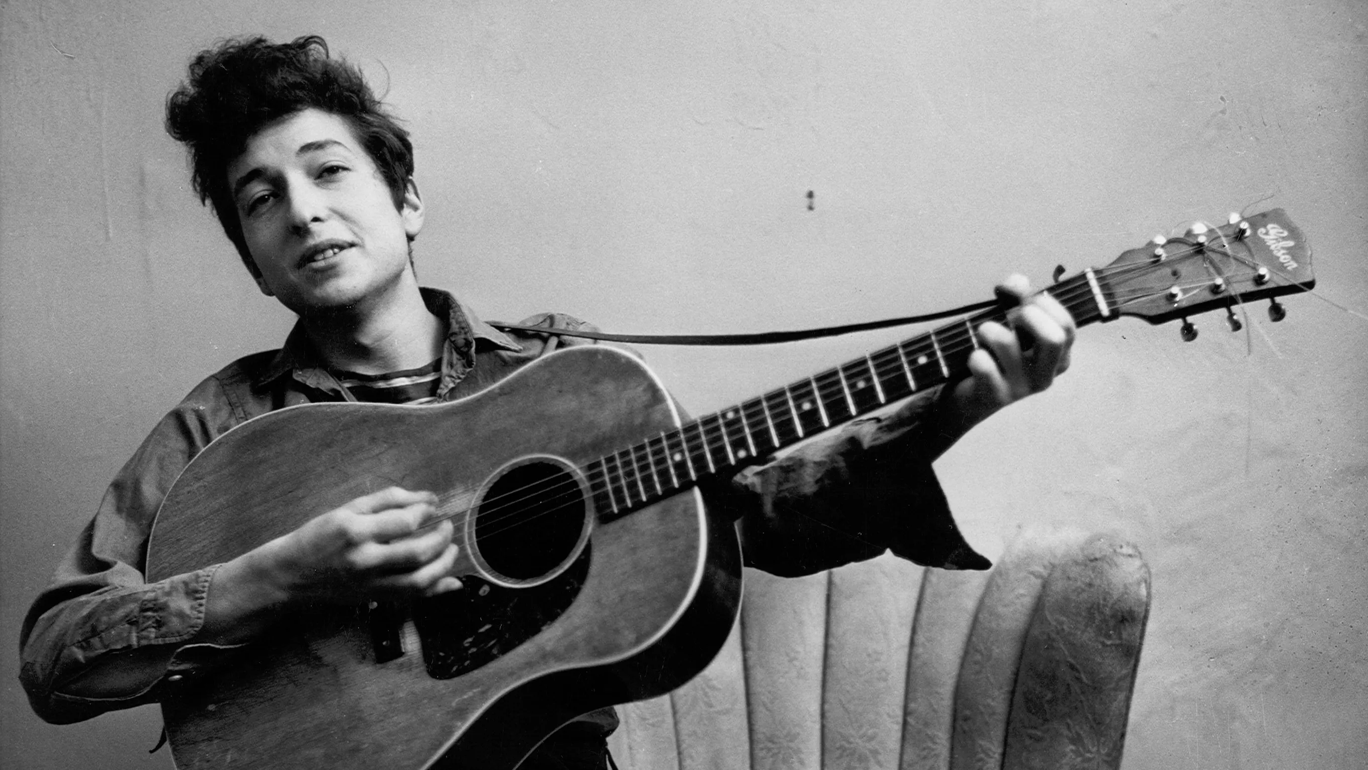 A young man with tousled hair is playing an acoustic guitar while seated, smiling slightly at the camera, against a plain background.