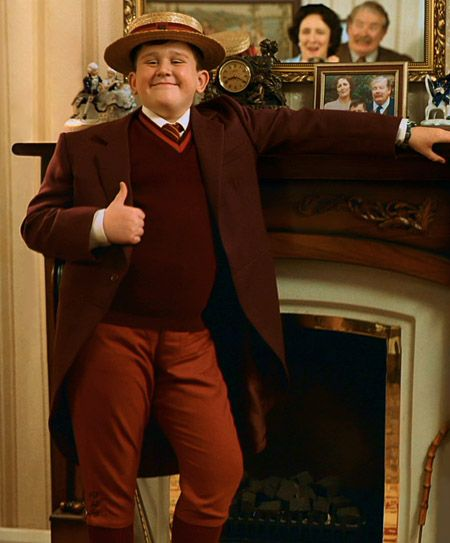 A young boy in a vintage outfit, including a maroon suit and a straw boater hat, strikes a confident pose with a thumbs-up gesture next to a fireplace decorated with family photographs.