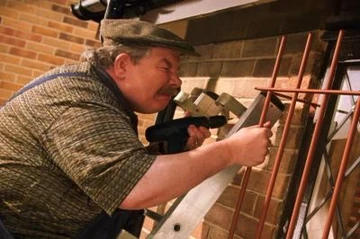 A man with a mustache, wearing a cap and patterned shirt, is using a power drill to secure metal bars to a brick wall while standing on a ladder.