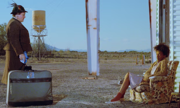 A man in a dark suit and hat stands with a suitcase, looking at a woman sitting in a floral armchair in a desolate landscape with sparse vegetation and a water tower in the background.