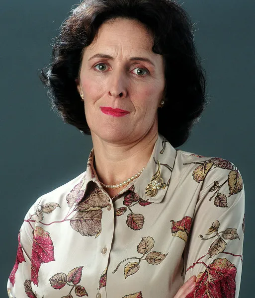 Portrait of a woman with dark, wavy hair, wearing a floral-patterned blouse and a pearl necklace, with a serious expression against a plain background.