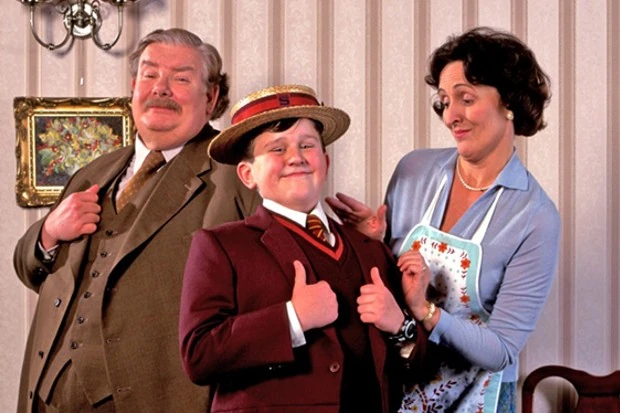 A cheerful young boy in a school uniform stands in the center, giving thumbs up. He is flanked by two adults: a man in a suit with a mustache on the left and a woman in an apron on the right, both smiling at the boy. The background features a patterned wallpaper and a framed picture.