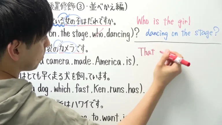 A person is writing on a whiteboard filled with English and Japanese sentences, while the individual is partially visible from behind, wearing a gray hoodie.