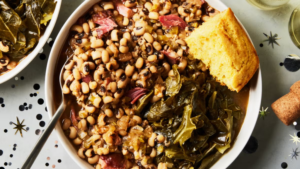 A bowl of black-eyed peas with ham, served with collard greens and a piece of cornbread, on a festive table decorated with confetti.