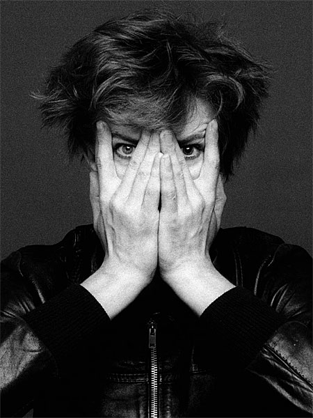 A person with tousled hair obscures their face with their hands, wearing a black leather jacket, set against a dark background.