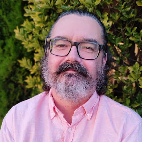 A man with a thick beard and glasses, wearing a light pink shirt, smiles softly against a backdrop of green foliage.