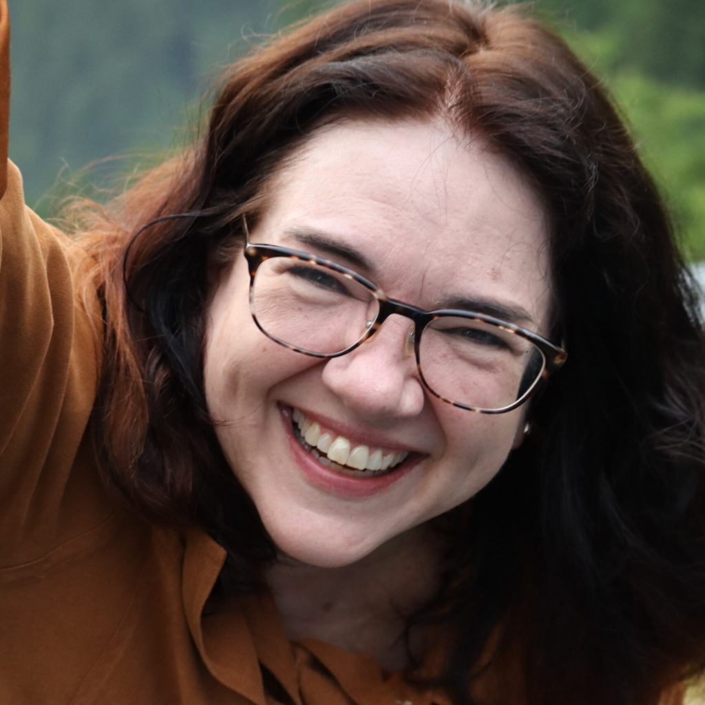 A woman with glasses and long brown hair, smiling joyfully with her head tilted slightly, wearing a brown top against a blurred green background.